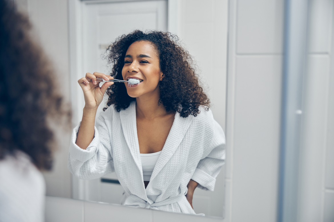 Black woman brushing her teeth in the mirror wearing white robe